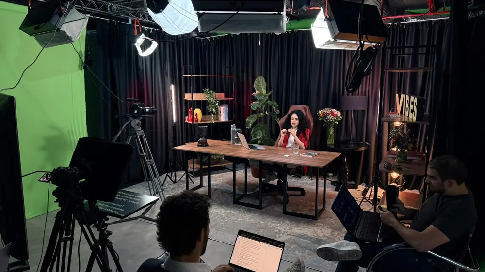 Gravação de curso no estúdio da Produtora 76: apresentadora à mesa, teleprompter e câmeras em primeiro plano, fundo verde à esquerda e equipe com notebooks acompanhando