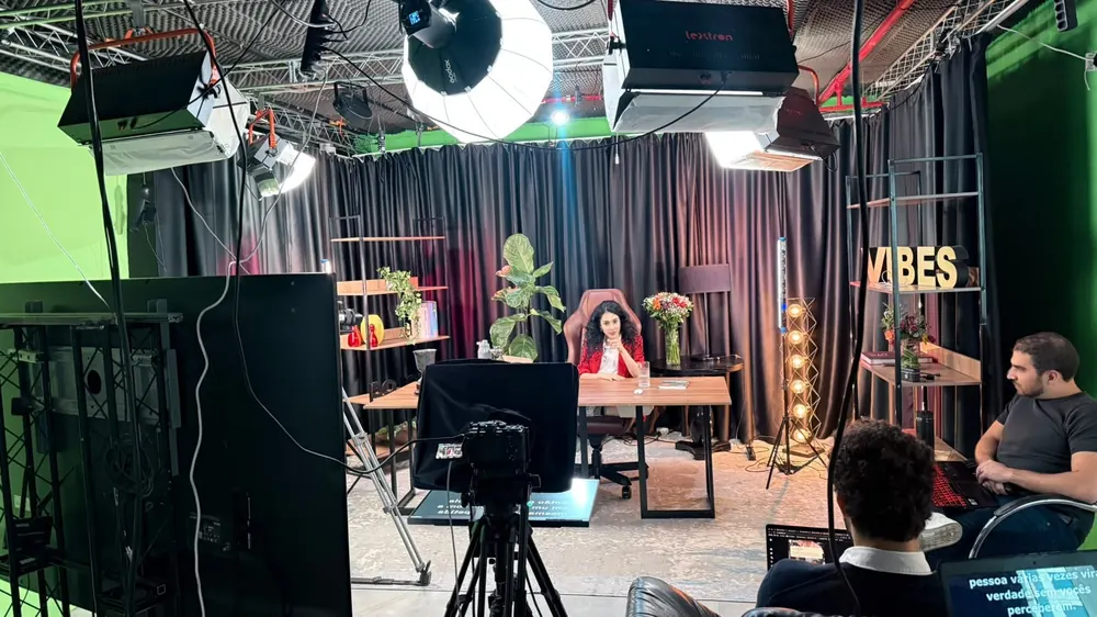 Câmera com teleprompter apontada para a apresentadora, iluminação de estúdio no teto e equipe acompanhando a gravação